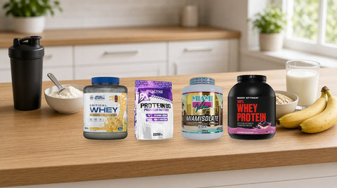 Proteine pudră Applied Nutrition Critical Whey, ProActive Protein 80, Miami Vibes Miamisolate și Body Attack 100% Whey pe un blat de lemn, lângă un shaker negru, un bol cu pudră, un pahar cu lapte și banane