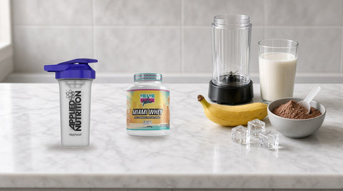 Shaker Applied Nutrition și o cutie de proteină Miami Whey pe un blat de marmură, lângă un blender mic, un pahar cu lapte, o banană, cuburi de gheață și un bol cu pudră proteică de ciocolată